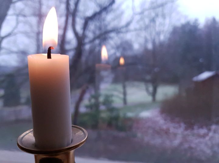 Valkoinen kynttilä metallisessa kynttilänjalassa ikkunalaudalla, kynttilän liekki heijastuu ikkunasta. Taustalla maalaismainen pihamaisema, maassa hieman lunta.