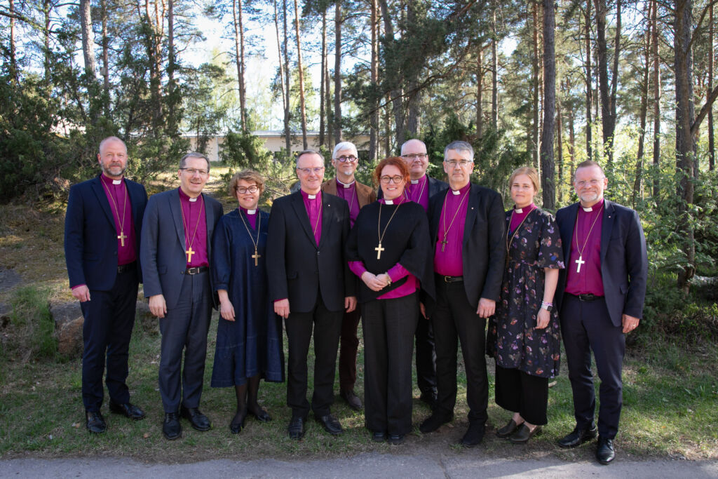 Ryhmä kirkon johtajia seisoo metsäisessä ympäristössä, pukeutuneina virallisiin vaatteisiin ja ristiriipuksiin.