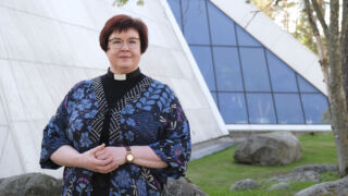 Sairaalapastori Tiina Peippo kukkakuvioisessa sinisävyisessä jakussa ulkona Hyvinkään kirkon edessä.