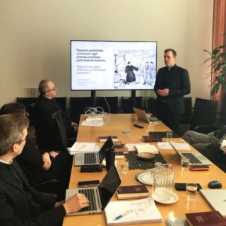 Ylemmän pastoraalitutkinnon keskustelu käynnissä tuomiokapitulin istuntosallissa. Pitkän pöydän ympärillä ihmisiä papinpuvuissa kuuntelemassa keskustelua.